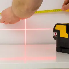Poziomica laserowa emituje czerwone linie krzyżujące się na białej powierzchni, obok dłoni trzymającej miarkę.