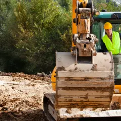 Operator koparki w żółtym kasku i kamizelce odblaskowej siedzi w kabinie maszyny na placu budowy.