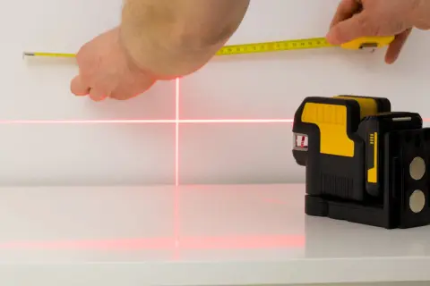 Poziomica laserowa emituje czerwone linie krzyżujące się na białej powierzchni, obok dłoni trzymającej miarkę.