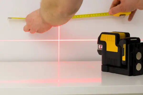 Poziomica laserowa emituje czerwone linie krzyżujące się na białej powierzchni, obok dłoni trzymającej miarkę.